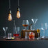 A bar-style still life with multiple clear drinking glasses and cocktail glassware arranged on a dark tabletop against a blue-gray background. Two hanging Edison bulbs illuminate the scene, with drinks, straws, a jigger, flowers in a vase, and wooden blocks used as styling props.