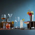 A bar-style still life with multiple clear drinking glasses and cocktail glassware arranged on a dark tabletop against a blue-gray background. Two hanging Edison bulbs illuminate the scene, with drinks, straws, a jigger, flowers in a vase, and wooden blocks used as styling props.