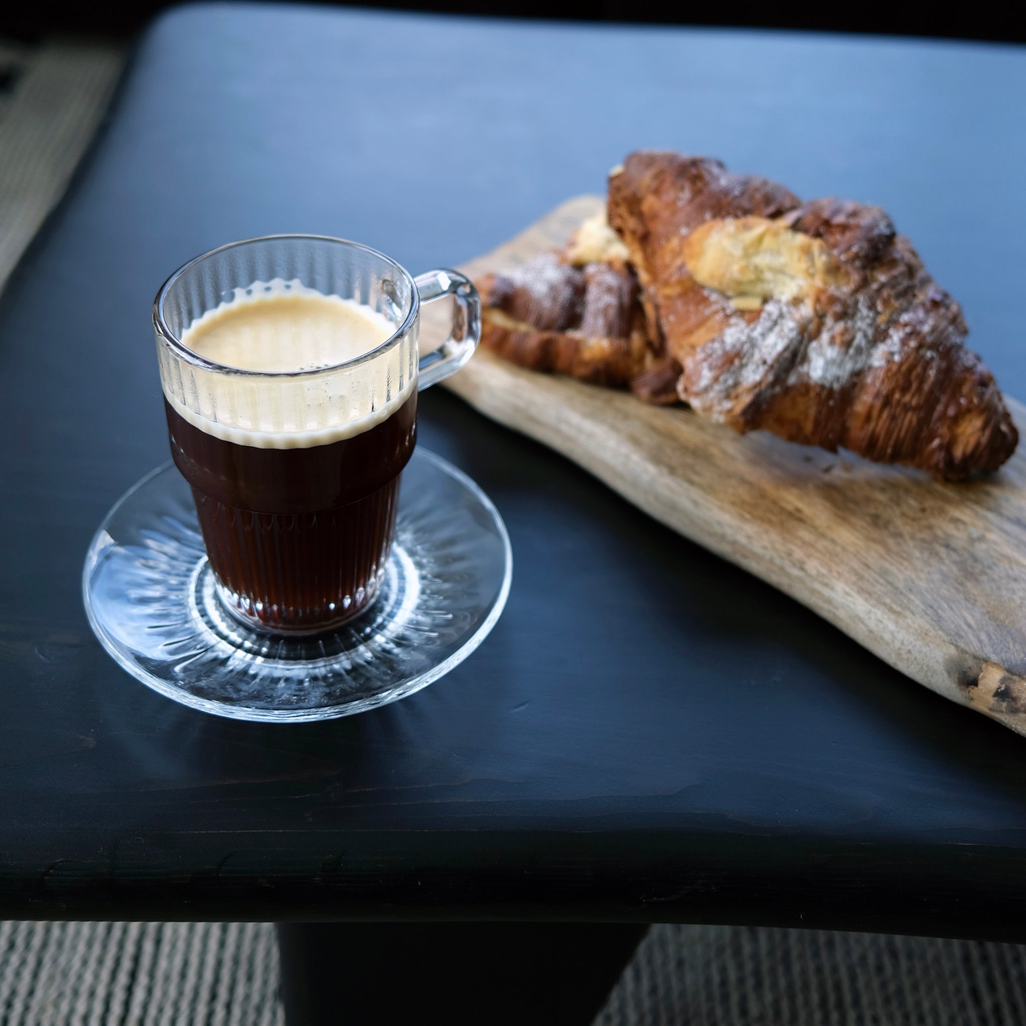 Glass of espresso on a dark surface with a wooden board of pastries.