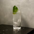 Clear Exclusiva Tom Collins glass with a clear cocktail and garnished with a green leaf on a black marbled surface and gray background