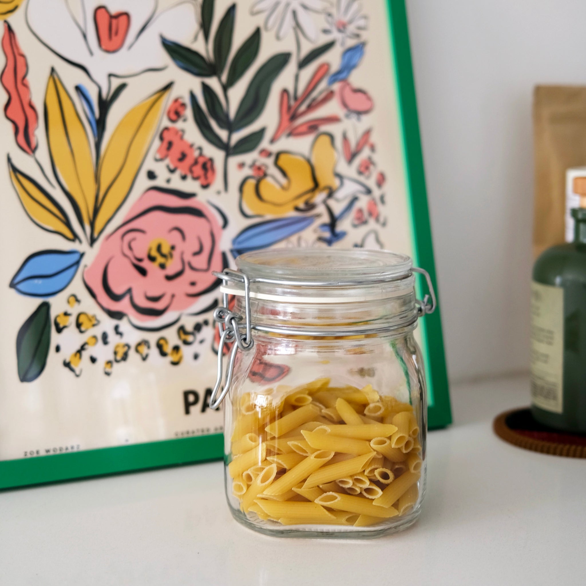 Clear glass jar with pasta on a table with a floral painting in the background