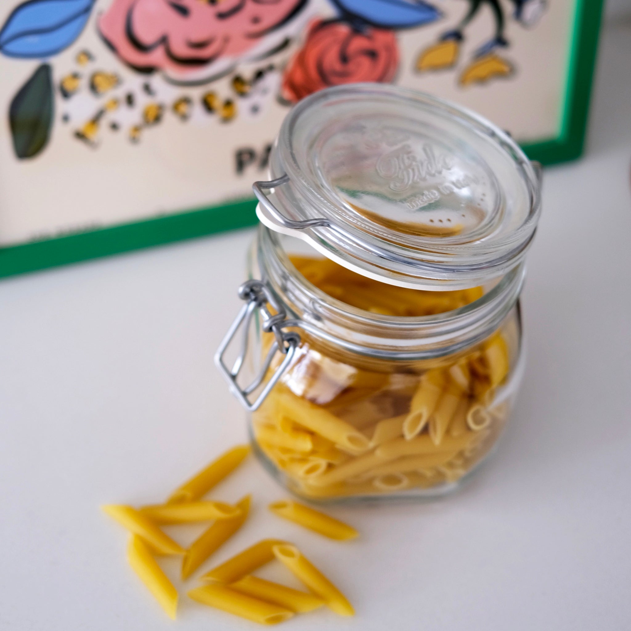 Glass jar with penne pasta on a white surface with a colorful box in the background