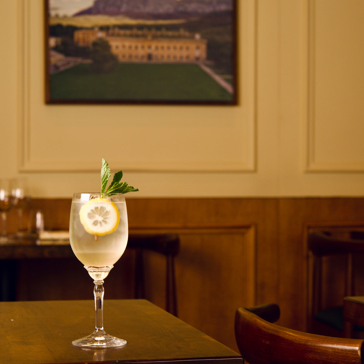 Florian White Wine glass filled with lemony cocktail and garnished with a slice of lemon and mint leaf on a table in a cozy room.